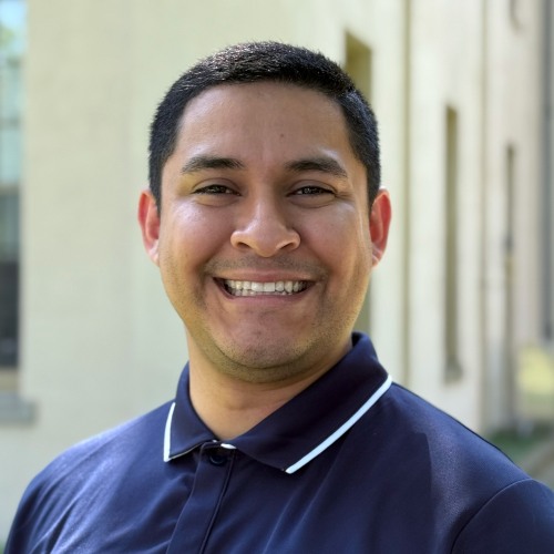 Headshot of Joshua Santos wearing a navy blue Brown University polo shirt, standing outside of a building